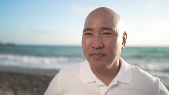 Middle Aged Asian Positive Bald Man Looking at Camera Smiling with Turquoise Mediterranean Sea Waves