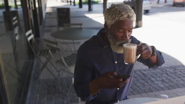 African american senior man using smartphone while drinking coffee sitting outdoors at cafe