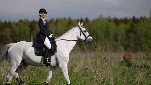 a Blonde in a Jockey Suit and a Riding Helmet Rides a White Horse Through a Field Against the