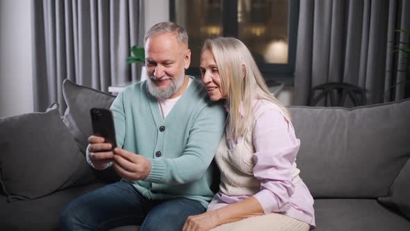 Elderly Couple Sits on a Sofa in the Living Room and View Photos on a Smartphone Family Day Positive