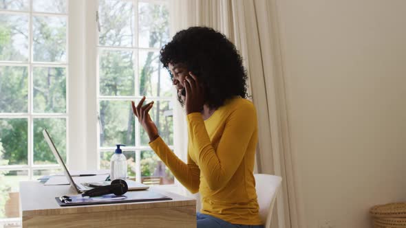 African american woman using laptop and talking on smartphone while sitting on her desk at home