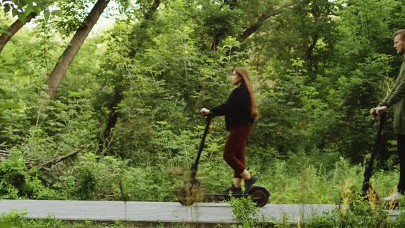 Woman and Man Drive Along Path in Park on Electric Scooters Side View