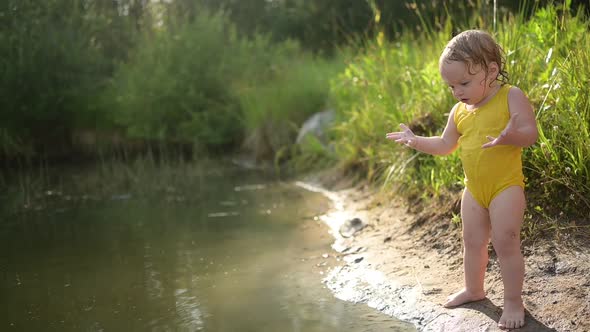 Little Funny Cute Blonde Girl Child Toddler in Yellow Wet Bodysuit Playing By the Lake Waterside