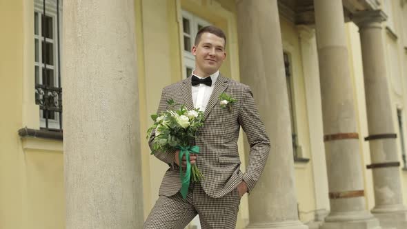 Groom, Young Man Stay Near House with Columns with Wedding Bouquet. He Waiting for His Beloved Bride