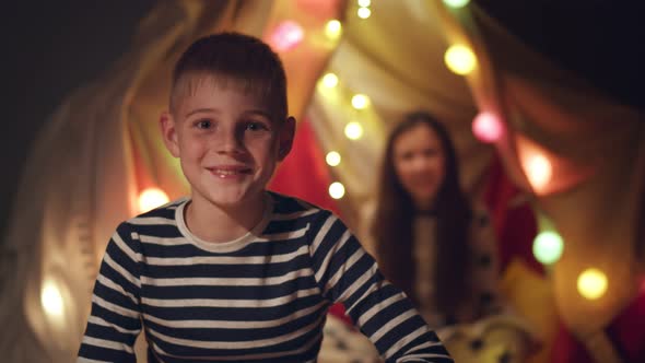 Happy Smiling Preteen Boy in Pajama Smiling at Camera with Sister Sitting in Wigwam on Background