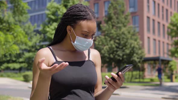 A Young Black Woman in a Face Mask Looks at a Smartphone and Gets Upset - an Office Building