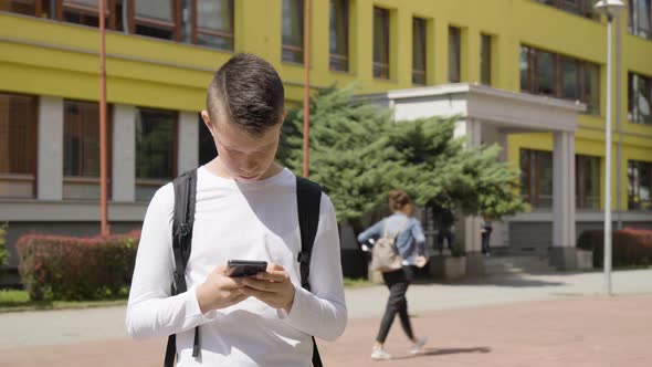 A Caucasian Teenage Boy Works on a Smartphone  a School in the Background