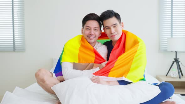 Attractive male lgbt couple sit on bed in bedroom in morning and looking at camera with LGBT flag.