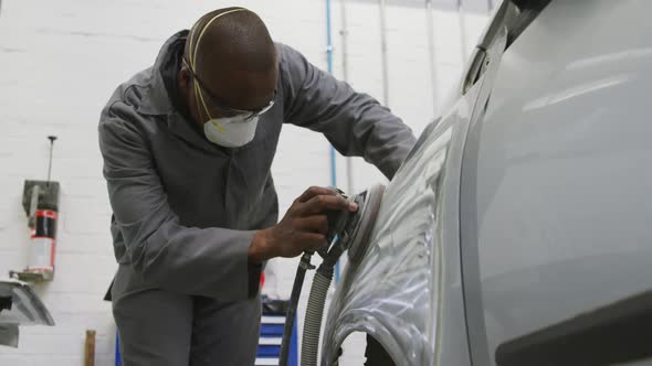 African American male car mechanic wearing a face mask and polishing a side of a car with a grinder