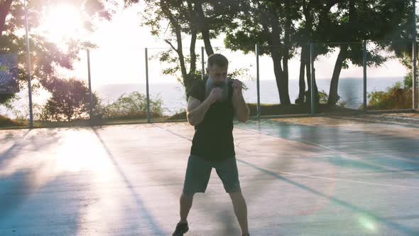 Middle Aged Man with Long Gray Beard Boxing with Shadow on Basketball Court During Sunrise