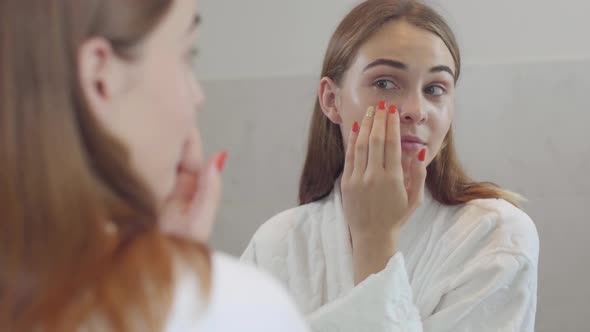 Attractive Young Woman with Different Colored Eyes Looking in the Mirror Applying the Daily Cream on