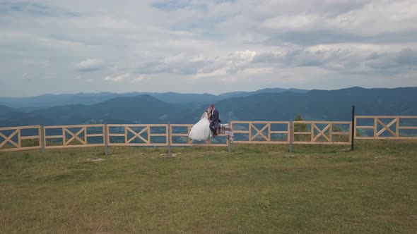 Groom with Bride Sit on the Fence Near Mountain Hills. Aerial Drone Shot