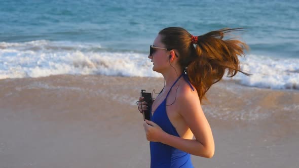 Young Woman in Swimsuit Running on Sea Beach and Listening Music. Girl Jogging Along Ocean Shore