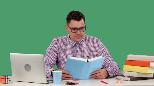 A Man in Glasses Sits at a Desk in Front of a Laptop and Reads a Book Lifts His Glasses and Smiles