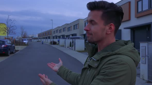 A Young, Handsome Man Stands on a Road and Gestures Approvingly at the Neighborhood Behind Him