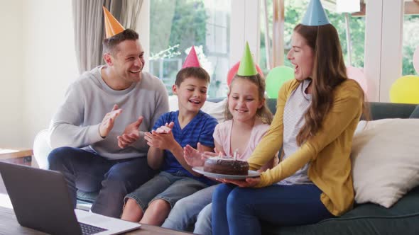 Caucasian woman in party hat holding birthday cake while family clapping and celebrating birthday at