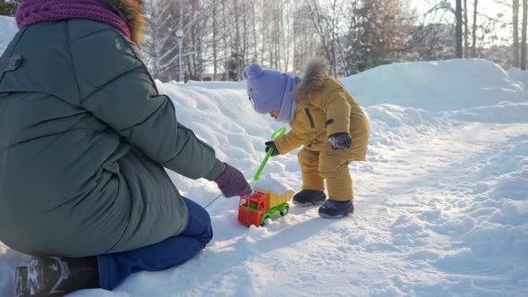 Mom plays with toddler in winter park with toys. Mom loads snow with her palms into a toy truck