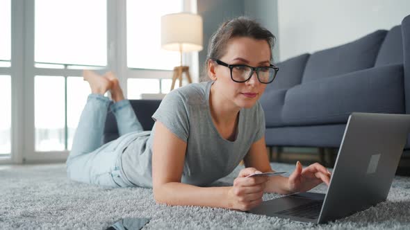 Woman with Glasses Is Lying on the Floor and Makes an Online Purchase Using a Credit Card and