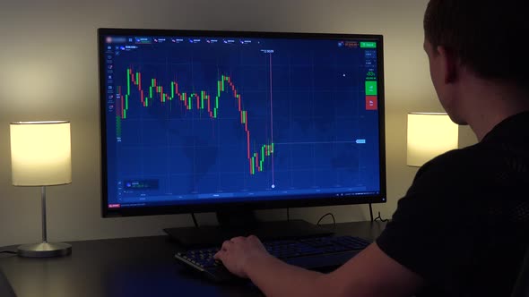 A Man Sits at a Table and Looks at a Candle Chart on a Computer Screen