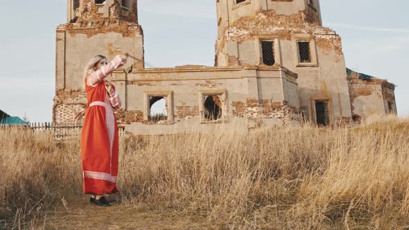 Woman in Red National Dress Wields Swords Against the Background of a Ruined Old Brick Church