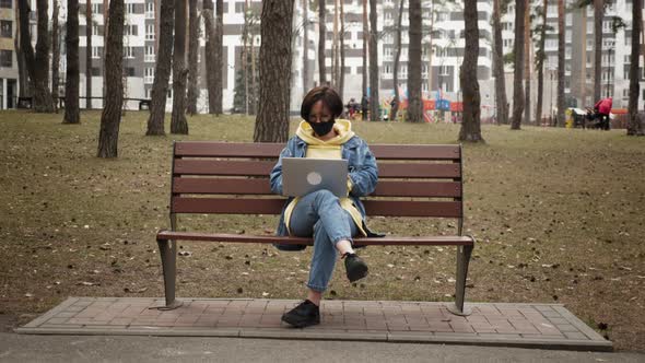 Woman in protective mask working in street with laptop sitting on bench in park.