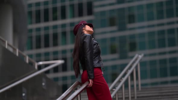 a Beautiful Dark-haired Female Model in a Purple Cap and Leather Jacket, in a Red Dress Poses in a