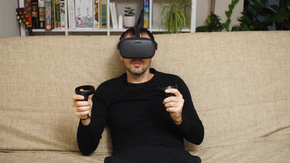 A bearded man in a VR helmet sits on a couch. In his hands are controllers