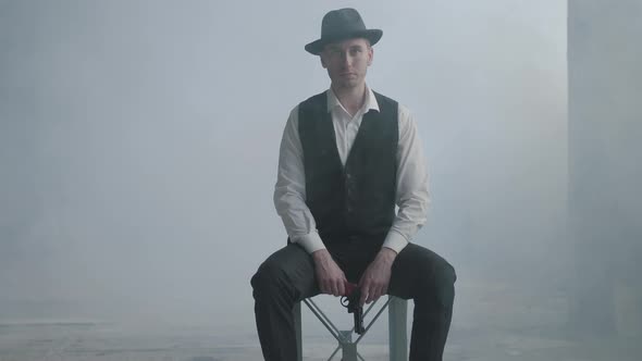 Handsome Confident Well-dressed Man in a Hat Sitting on the Chair Holding a Gun in the Smoky Room in