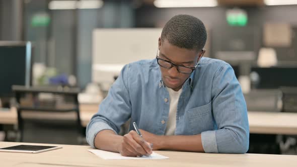 Hardworking Young African American Man Writing on Paper in Office