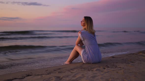 Backside View Young Woman with Naked Legs Sitting at Sea on the Beach Wearing Blue Summer Dress