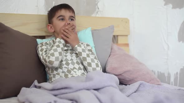 A Sick Little Boy Coughs While Sitting in Bed and Covers Himself with a Blanket