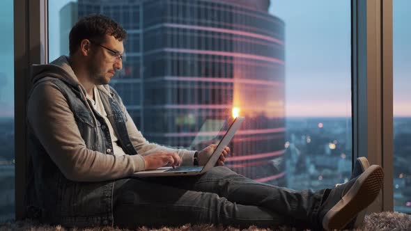 Man with a Laptop Is Sitting By the Window on a High Floor of a Skyscraper