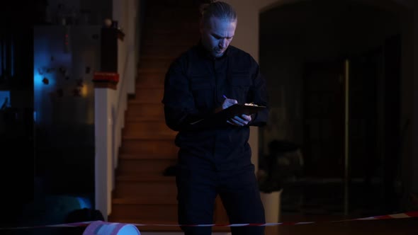 Portrait of Handsome Male Police Officer Filling in Report Standing in Darkness in Robbed House