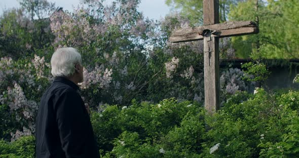 Sad Old Gray Haired Man Stands Near Big Old Wooden Cross at the Cemetery