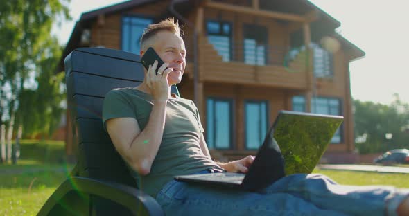 Happy Young Man Sitting on Background of Country House with Laptop and Talking on the Phone