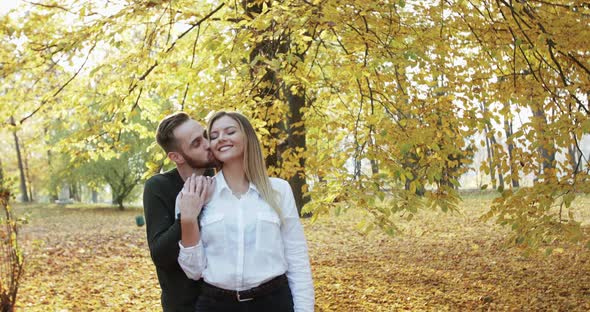 Lovely Man Embraces His Girl and Kisses Her in Neck in Autumn Park