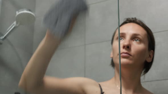 Woman wiping glass door of bathtub in bathroom.