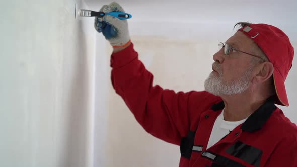 Mature Graybearded Man in a Red Cap and Glasses Paints the Wall with a White Paintbrush in His New