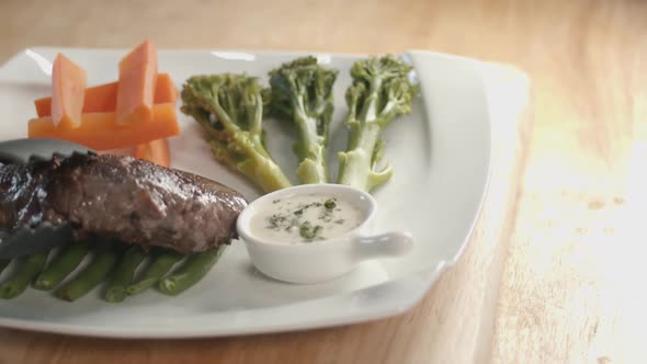 Slider Shot of a Square Plate With Vegetable on it Then the Serving of Steak Next to the Sauce Pot o