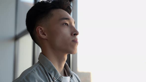 Young Asian Man Stands in Office Looking at Panoramic Window