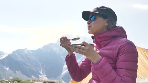 A Young Woman Traveler in a Pink Down Jacket is Having Breakfast with Porridge in the Mountains Near