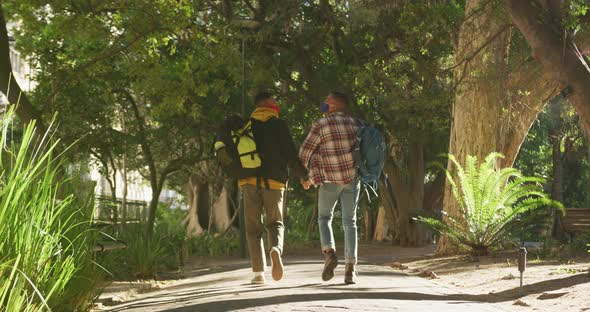 Back view of two happy mixed race gay male couple walking and talking in park with backpacks