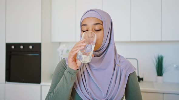 Young Arab Woman in Hijab Drinks Water From a Glass While Standing in the Kitchen at Home