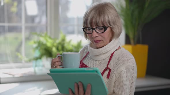 Concentrated Absorbed Caucasian Senior Woman Reading News on Tablet Standing with Coffee Cup in the