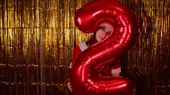 Woman in Santa Costume Holds Red Figure Two on Background of Shimmering Golden Tinsel