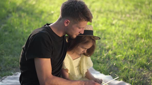 Side View Young Brunette Man and Cute Girl Sitting on Sunny Park Lawn Surfing Internet on Tablet