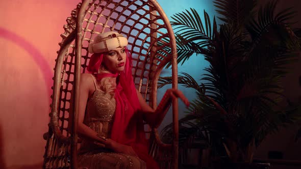 a Woman in a Turban with a Red Scarf and a Sequined Dress is Sitting in a Wicker Chair