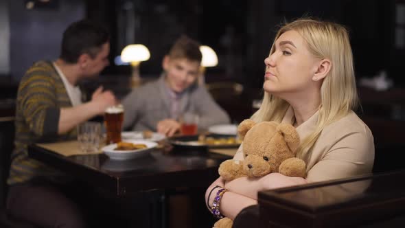 Sad Caucasian Woman Posing with Teddy Bear in Restaurant As Husband and Son Talking at Background