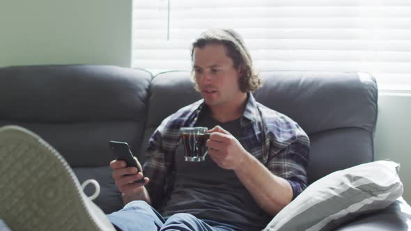Happy caucasian man sitting on sofa in living room using smartphone and drinking coffee