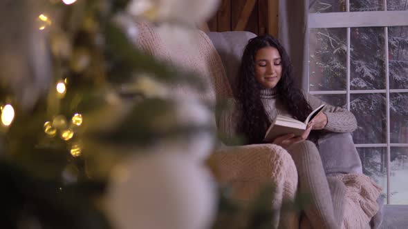 A Girl Sits in a Chair Near the Christmas Tree and Reads a Book